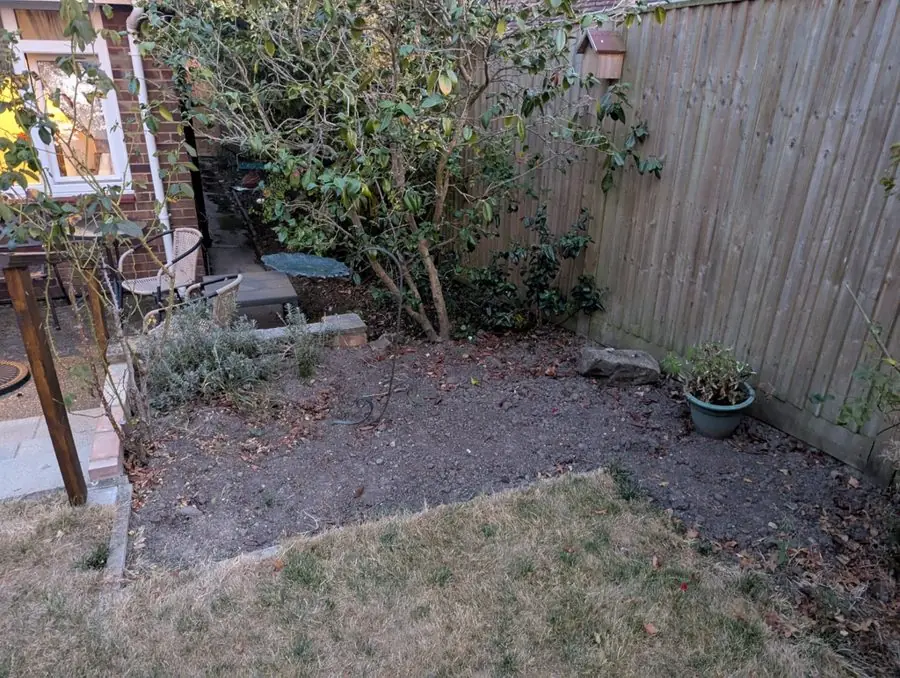 A bare corner of dry soil by a fence and a small patio, before the redesign