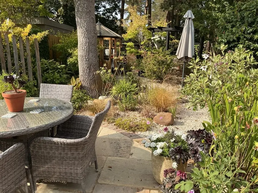 A patio table and chairs beside gravel beds under a tall tree, before the redesign