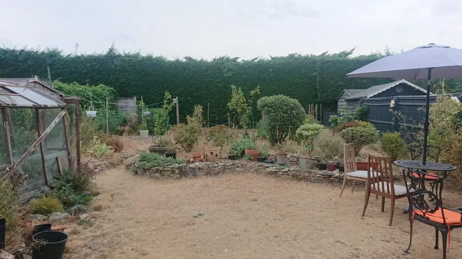 A dusty bare yard with a parasol, table, chairs and a greenhouse, before the redesign